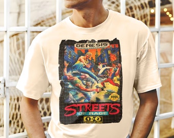 Puede incluir: Una camiseta blanca con un diseño vintage de cartucho de videojuego Sega Genesis. El diseño presenta el juego "Streets of Rage" con una imagen pixelada de los personajes del juego luchando en una calle de la ciudad. El texto "Sega Genesis" y "Streets of Rage" está impreso en rojo y negro.