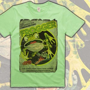Puede incluir: Camiseta verde claro con un gráfico desgastado del videojuego clásico Frogger. El gráfico incluye una gran rana verde, el título del juego y texto que indica que es un cartucho de Parker Brothers para los sistemas de videojuegos Atari y Sears.