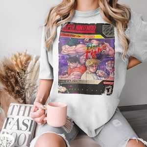 Può includere: Una maglietta bianca con un design di cartuccia di gioco Super Nintendo vintage con il testo "Final Fight" e una grafica di tre personaggi che combattono. La cartuccia di gioco è circondata da un bordo nero con testo bianco.