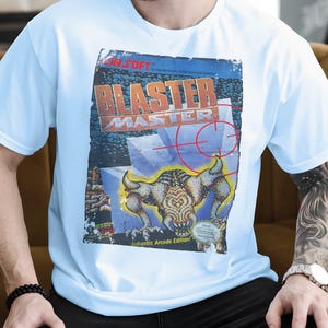 Könnte beinhalten: Hellblaues T-Shirt mit dem Cover-Artwork des Videospiels Blaster Master. Das Design zeigt den Titel des Spiels in Orange und eine Kreaturenillustration. Das T-Shirt hat einen klassischen Rundhalsausschnitt.