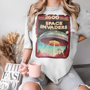 Puede incluir: Una camiseta blanca con un gráfico vintage de cartucho de videojuego Atari 2600 Space Invaders. El gráfico presenta un esquema de color rojo, blanco y azul con una nave espacial y estrellas.