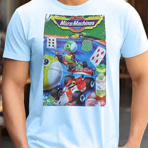 May include: Pale blue t-shirt with a vibrant Micro Machines graphic. The design includes miniature race cars, a pool ball, playing cards, and the Micro Machines logo. The overall theme is retro toys.