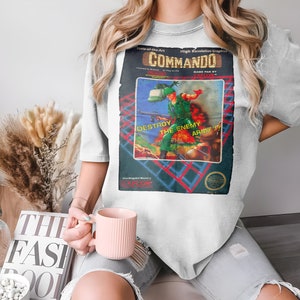 Pode incluir: Uma camiseta branca com uma estampa gráfica de videogame vintage do jogo Commando. O gráfico apresenta um soldado segurando uma arma e o texto "DESTROY THE ENEMY ARMY!" em vermelho e azul.