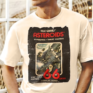 Puede incluir: Una camiseta blanca con un gráfico de estilo vintage del juego Atari Asteroids. El gráfico presenta el título del juego, una nave espacial y el texto "TELE-GAMES", "HYPERSPACE THRUST CONTROL", "FOR 1 OR 2 PLAYERS", "CONTAINS ONE CARTRIDGE" y "GAMES".