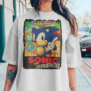 May include: A white t-shirt with a vintage-style graphic of Sonic the Hedgehog. The graphic features Sonic in his classic blue and red design, with the text "Genesis 16-bit Cartridge" and "Sonic the Hedgehog" in a retro font.