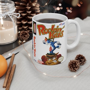 May include: White ceramic mug with a colorful illustration of a video game character from the game "Rockin' Kats" on the front. The mug has a black handle and is filled with a dark liquid.