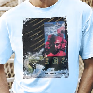 May include: Pale blue t-shirt with a graphic print. The print features a dragon, three figures, and Japanese text. The design has a vintage, distressed aesthetic with a black and gold border. The t-shirt is a solid pale blue colour.