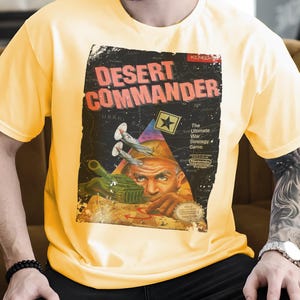 Puede incluir: Camiseta amarilla con un gráfico retro de "Desert Commander", un juego de estrategia de guerra. El diseño incluye la cara de un soldado, un tanque y aviones de combate, con el logotipo de Nintendo. La camiseta es de un material suave.