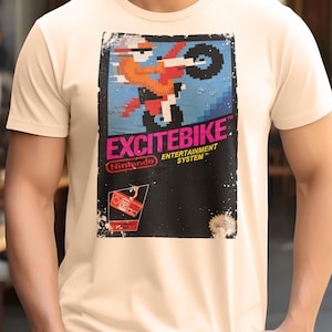 May include: A cream-colored t-shirt featuring a retro video game graphic. The design showcases a pixelated motocross racer in orange and black, with the word "EXCITEBIKE" in pink and the Nintendo logo. The shirt has a classic crew neck.
