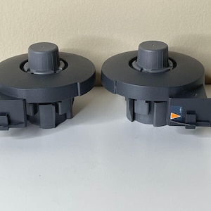 Puede incluir: Dos componentes de plástico gris con un diseño circular y una perilla central. Cada componente tiene una sección rectangular en el lateral con un triángulo naranja y blanco. Los artículos están sobre una superficie blanca.