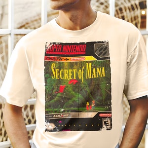 May include: A white t-shirt with a vintage Super Nintendo game cartridge design featuring the text "Secret of Mana" and a jungle scene. The design is faded and worn, giving it a retro look.