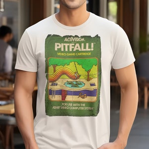 Puede incluir: Una camiseta blanca con un gráfico de estilo vintage del videojuego de Atari Pitfall! El gráfico presenta una escena de jungla verde con un personaje balanceándose en una liana sobre un pozo de serpientes. El texto "Activision Presents Pitfall! Video Game Cartridge" está impreso en el gráfico.