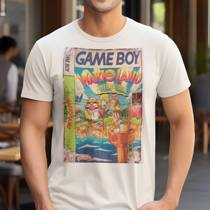Puede incluir: Camiseta blanca con un diseño de cartucho de Game Boy vintage con Wario Land: Super Mario Land. El diseño está en un estilo descolorido y desgastado.