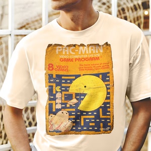 Peut inclure: Un t-shirt blanc avec un design vintage de programme de jeu Pac-Man. Le design présente un personnage Pac-Man jaune dans un labyrinthe bleu avec des fantômes. Le texte sur le design indique "PAC-MAN VIDEO COMPUTER SYSTEM GAME PROGRAM 8 VIDEO GAMES The home version of one of your favourite arcade games ONE PLAYER TWO PLAYERS 02646".