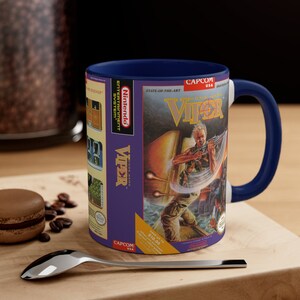 Pode incluir: Uma caneca de cerâmica azul com interior branco que apresenta um design de cartucho de videogame vintage. A frente da caneca mostra a capa do jogo Nintendo Entertainment System "VIPER" da Capcom. O texto no cartucho diz "STATE-OF-THE-ART", "VIPER", "CODE NAME", "CAPCOM USA", e "SAVE UP TO 10,00 €".