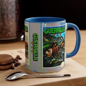 May include: A blue and gray ceramic mug with a design of a video game cartridge for the game "Guerrilla War" on the front. The game cartridge is a light gray color with a green and yellow image of a jungle scene with soldiers. The text "Guerrilla War" is printed in white on the cartridge. The mug has a blue handle.