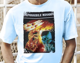 May include: Light blue t-shirt featuring a graphic from "Impossible Mission." The design includes two figures, a fiery explosion, and a futuristic vehicle. The text "IMPOSSIBLE MISSION" is at the top of the graphic.