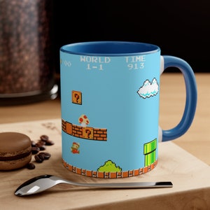 Könnte beinhalten: Eine blau-weiße Keramik-Tasse mit einer pixeligen Szene aus dem klassischen Videospiel Super Mario Bros. Die Tasse hat einen blauen Innenraum und Henkel, und das Design zeigt Mario, einen Pilz und den Text "WORLD 1-1 TIME 913."