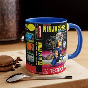 Pode incluir: Uma caneca de cerâmica azul com um gráfico do jogo Ninja Gaiden II: The Dark Sword of Chaos para Nintendo Entertainment System. O título do jogo está em japonês e inglês. A caneca tem uma alça azul.