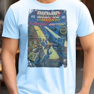 May include: Pale blue t-shirt with a retro comic book design for "Bionic Commando" by Capcom. The graphic shows action scenes with robots and soldiers, with the game title in bold red and blue lettering.