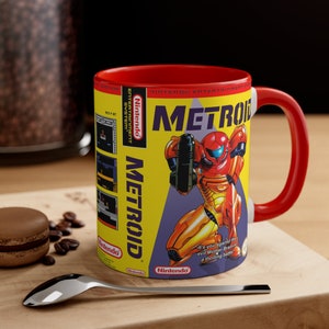 May include: A ceramic coffee mug featuring the video game Metroid. The mug has a yellow background with the game's logo and a character illustration. The interior and handle are red. The mug also has the Nintendo logo.