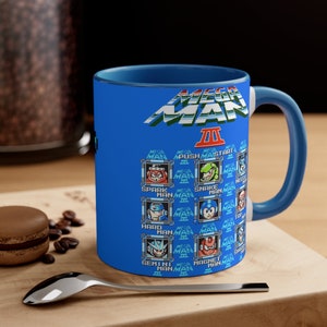 May include: A blue ceramic mug featuring a graphic of Mega Man 3. The mug displays the title "Mega Man 3" and the names of the eight robot masters from the game: Spark Man, Snake Man, Hard Man, Top Man, Gemini Man, Magnet Man, and the text "Mega Man 3" repeated multiple times.