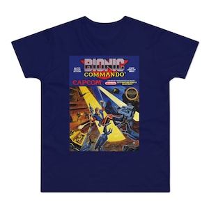 May include: A blue t-shirt with a retro video game box design featuring a character in a blue and red suit jumping from a platform. The box art reads "Bionic Commando" and "Capcom".