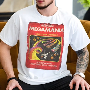 Puede incluir: Camiseta blanca con un gráfico de estilo vintage de un cartucho de videojuego Atari. El gráfico presenta el texto "ACTIVISION. MEGAMANIA VIDEO GAME CARTRIDGE" y una ilustración colorida de una nave espacial volando a través de un rastro de arcoíris.