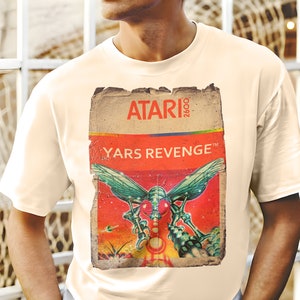 Puede incluir: Una camiseta blanca con un diseño de cartucho de juego Atari 2600 vintage con el texto "Yars Revenge" y un gráfico de una nave espacial.