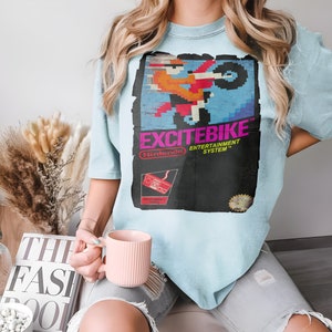 Pode incluir: Uma camiseta azul claro com um gráfico do jogo "Excitebike" do Nintendo Entertainment System. O gráfico está em um estilo pixelado retrô e apresenta uma pessoa pilotando uma motocicleta.
