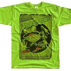 Puede incluir: Una camiseta verde lima con un gráfico desgastado del videojuego clásico Frogger. El gráfico incluye una rana verde, un cocodrilo y el título del juego. El texto en la camiseta dice "VIDEO GAME CARTRIDGE FROM PARKER BROTHERS" y "FOR ATARI & SEARS VIDEO GAME SYSTEMS."