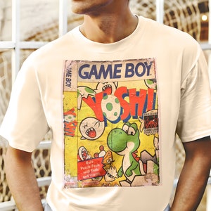 Op de afbeelding: Een wit T-shirt met een vintage Game Boy-cartridge ontwerp met Yoshi. De cartridge is geel met rode, groene en blauwe accenten. De tekst op de cartridge luidt "GAME BOY" en "Rare Puzzle Panic with Yoshi and Friends".