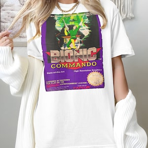 Könnte beinhalten: Weißes T-Shirt mit einer lila und grünen Grafik einer Videospielkassette für Bionic Commando. Der Text auf der Grafik lautet "Bionic Commando", "State-of-the-Art", "High Resolution Graphics", "Licensed by Nintendo for play on the Entertainment System", "Game Pak of Capcom".
