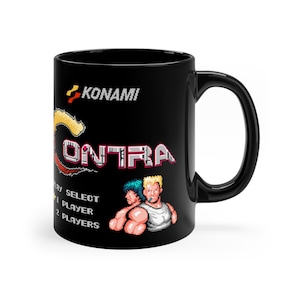 Op de afbeelding: Zwarte keramische mok met een wit en rood "KONAMI" logo en het woord "CONTRA" in een pixellettertype. De mok heeft een pixelafbeelding van twee personages uit het videospel "Contra" met de tekst "PLAY SELECT 1 PLAYER 2 PLAYERS".