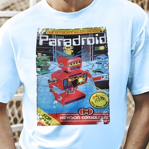 May include: Light blue t-shirt featuring a retro graphic of a red robot with the text "Paradroid" and "The Ultimate Brain Drain!". The design includes other robots and a gold medal award. The shirt is for ages 6 and up.