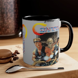 Puede incluir: Taza de cerámica blanca con interior y asa negros. La taza presenta la palabra "CONTRA" en una fuente retro, junto con un gráfico de dos soldados y un alienígena. El logotipo de Konami es visible.