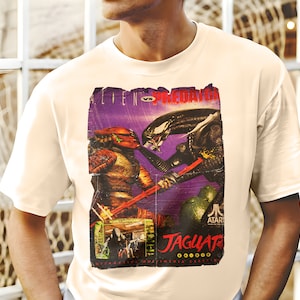 Könnte beinhalten: Weißes T-Shirt mit einem Grafikdruck im Vintage-Stil des Atari Jaguar Videospiels 'Alien vs Predator'. Die Grafik zeigt die Charaktere Predator und Alien in einer Kampfszene.