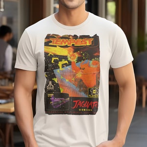 Puede incluir: Camiseta blanca con un gráfico de estilo vintage del juego de arcade Tempest. El gráfico presenta un monstruo rojo y naranja con el texto "Tempest" y "Atari" en el diseño.