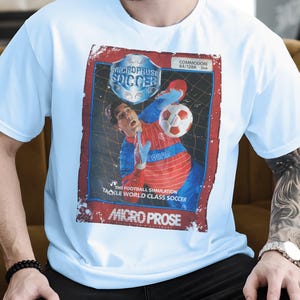 Könnte beinhalten: Ein hellblaues T-Shirt mit einem Retro-Grafikdesign für "Microprose Soccer". Das Design zeigt einen Fußballspieler, einen Ball und den Text "Commodore 64/128K". Das Shirt hat auch den Text "The Football Simulation Tackle World Class Soccer".