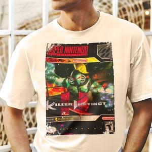 Op de afbeelding: Een wit T-shirt met een vintage-stijl afbeelding van een Super Nintendo-spelcartridge voor Killer Instinct. De afbeelding toont een groen en rood personage met een helm en een zwaard. De tekst op de cartridge luidt "Super Nintendo", "Only For", "Nintendo", "Killer Instinct", "Rareware" en "Nintendo".