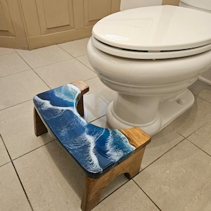 May include: A wooden bathroom step stool with a blue and white resin top featuring a wave design. The stool is in a bathroom setting next to a white toilet.