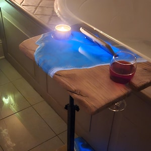 May include: A wooden bath tray with a blue resin design, holding a lit candle and a glass of red wine. The tray is supported by a black metal stand. The word "MOMENT" is in the background.