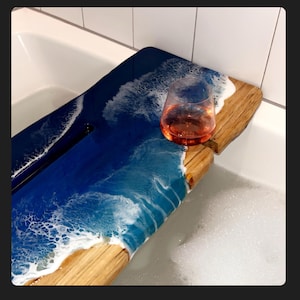 Peut inclure: Un plateau de bain en bois avec un design d'onde océanique en résine bleue et blanche. Le plateau a un porte-téléphone intégré et un verre de vin repose sur le plateau.