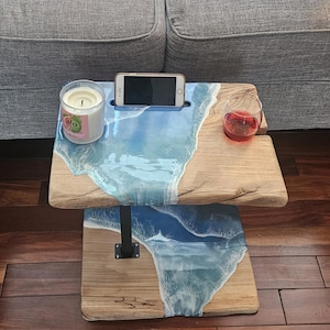 May include: A two-tiered wooden side table with a blue and white epoxy resin top. The top tier has a phone slot, a glass of red wine, and a candle. The bottom tier has a blue and white epoxy resin wave design.