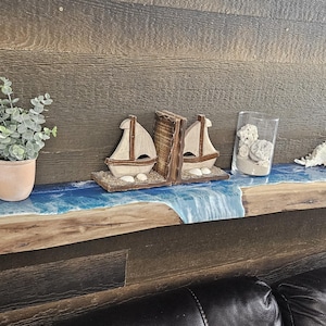 Peut inclure: Une étagère en bois avec un design de vague en résine bleue. L'étagère est montée sur un mur en bois foncé et décorée de petits voiliers en bois, d'une plante en pot et de coquillages.