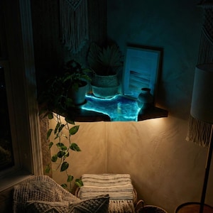Peut inclure: Une étagère flottante en bois avec un motif de résine bleu brillant, éclairée par une lumière chaude. L'étagère est décorée de plantes, d'une image encadrée et d'un petit vase. La pièce a une esthétique bohème et confortable.