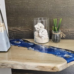 May include: A wooden shelf with a blue resin river flowing through it. The shelf is decorated with a glass bottle filled with sand and seashells, a glass cylinder with sand and coral, and a small potted succulent.