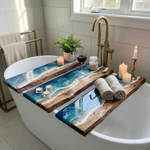 May include: Three wooden bath caddies with a resin ocean design, resting on a white bathtub. The caddies hold candles, rolled towels, a plant, and a glass of wine. The bathroom has a gold faucet and towel rack.