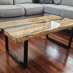 May include: A wooden coffee table with a live edge top and a black epoxy river running through the center. The table has a clear epoxy finish and sits on a black metal frame.