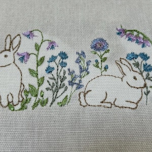 May include: Embroidered artwork depicting two white rabbits amongst blue and purple wildflowers. The rabbits are outlined in brown thread, and the flowers have green stems and leaves. The background is a light beige fabric.